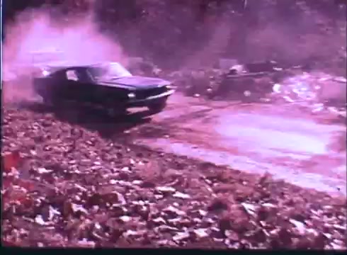 The still features a black car speeding through a rugged, outdoor setting. Dust and debris are kicked up behind the vehicle, suggesting high speed and action. The environment appears to be littered with leaves and other materials, indicating a rural or wooded area. The overall tone is gritty, reflecting the thrilling and adventurous themes of the 'Last American Hero' trailer.