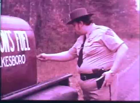The still depicts a law enforcement officer in a uniform, including a hat, interacting with an old fuel tank on a roadside. The tank prominently displays the words 'FUEL' and 'HICKSBORO.' The scene is set in a rural or wooded area, with muted colors typical of 16mm film, suggesting a vintage aesthetic that reflects the film's time period.