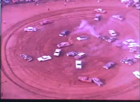 The still depicts a dynamic scene from the 'Last American Hero' trailer, showcasing a circular dirt racetrack filled with various cars. The vehicles appear to be engaged in a demolition derby or race, with some dust and smoke trails indicating action. The overall atmosphere conveys excitement and chaos typical of classic car racing films.