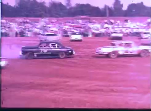 The still from the 'Last American Hero' trailer depicts a vibrant, dusty racetrack scene filled with spectators. Two cars are intensely racing, with one black car kicking up dirt as it maneuvers around another vehicle. The atmosphere captures the excitement and thrill of classic dirt track racing.