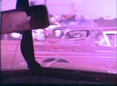 The still features a view from inside a car, showcasing a rearview mirror in focus with a hand visible. In the background, several cars, likely at a racetrack or a similar event, can be seen, creating a lively atmosphere. The color palette has a faded or vintage quality, typical of 16mm film, contributing to a nostalgic feel. This captures a moment related to racing, reflecting themes from 'Last American Hero.'