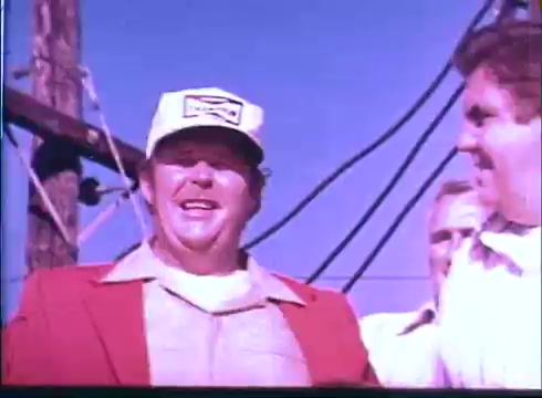 The still features a cheerful man in a white cap and a red jacket, smiling and engaging with others. He appears to be in a celebratory or informal setting, likely outdoors, with power lines in the background. The overall tone suggests a lighthearted moment, typical of sports-related or community themes found in the trailer for 'Last American Hero.'