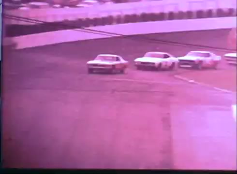 The still features a scene from the 'Last American Hero' trailer, showcasing a thrilling moment on a racetrack. Several cars, including a prominent white and red car, are in tight formation as they maneuver through a curve. The image has a vintage look typical of 16mm film, capturing the excitement of classic stock car racing.