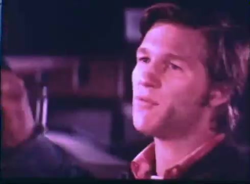 The still features a young man with light brown hair and a confident expression, dressed in a casual jacket over a collared shirt. The background is blurred, suggesting a nighttime scene, possibly in an outdoor or street setting. The lighting creates a nostalgic tone, capturing a moment of dialogue or contemplation.