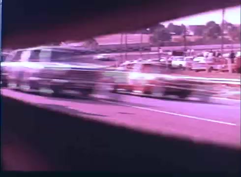 The still from the 'Last American Hero' trailer depicts a dynamic racing scene, showcasing blurred cars speeding along a road. The image conveys a sense of motion and excitement typical of a racing film, with spectators visible in the background. The color tone gives it a vintage feel, characteristic of 16mm film.