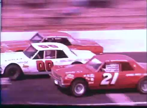 The still captures a thrilling moment from the 'Last American Hero' trailer, showcasing three race cars competing closely on a racetrack. The prominent vehicles are adorned with distinctive numbers: a white car with '08,' a vibrant red car marked '21,' and another red vehicle positioned behind them. The image conveys a sense of speed and competition, typical of stock car racing. The backdrop hints at a lively racing environment, emphasizing the excitement of the sport.