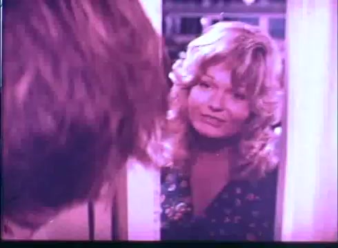 The still from the 'Last American Hero' trailer features a woman with wavy, shoulder-length hair, looking at someone off-screen with a pleasant expression. She appears to be standing in a doorway, suggesting an intimate or personal interaction. Her outfit is a colorful floral print, indicative of a warm, casual setting. The overall tone is nostalgic, fitting the film's themes.