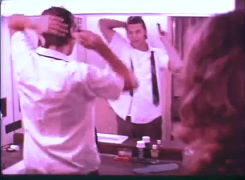 The still from the 'Last American Hero' trailer depicts a male character in a bathroom setting. He is adjusting his hair while facing a mirror, wearing a white shirt with a black tie and suspenders. A female figure is partially visible in the foreground, adding to the scene's intimate vibe. The lighting has a soft, warm quality, typical of the film's era, suggesting a moment of personal grooming and preparation.