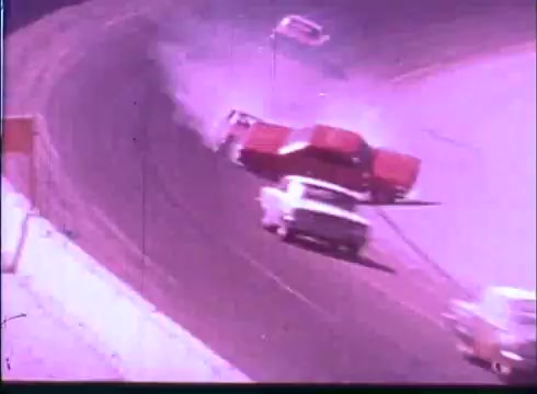 The still from the 'Last American Hero' trailer shows a dramatic moment on a racetrack. A red car is skidding out of control, possibly colliding with other vehicles, as tire smoke billows around it. The scene captures the intensity and excitement of racing, with multiple cars in motion and an air of chaos. The colors and film quality suggest a vintage look typical of 16mm footage.