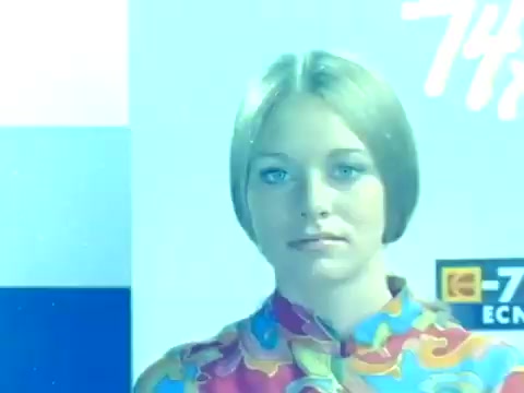The still from the trailer of 'When Women Lost Their Tails' features a young woman with a stylish bob haircut, dressed in a colorful patterned shirt. The image has a cool blue tint, possibly indicative of color grading or lighting effects used in the 1970s. The background includes signage or logos suggesting a modern or promotional context, fitting for the American release of the Italian film. This frame captures the film's aesthetic, reflecting the fashion and cultural sensibilities of the era.