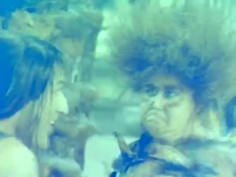 The still from the trailer of *When Women Lost Their Tails* features two characters engaged in a whimsical interaction, set against a vibrant, possibly natural backdrop. One character appears to have wild hair and a comical expression, suggesting a humorous or fantastical element typical of the film's genre. The overall color palette is bright and somewhat surreal, matching the playful tone of the movie, which is a comedic take on themes of gender and society from the early 1970s. This specific still highlights the film's blend of fantasy and humor, indicative of its appeal during its American release.
