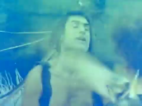 The still from the trailer of 'When Women Lost Their Tails' features a scene drenched in an ethereal blue hue, emphasizing a surreal or dreamy quality. It depicts a male character in a frantic or expressive moment, with his hair flowing and eyes closed, suggesting intense emotion or a dramatic action sequence. The background hints at an adventurous setting, possibly underwater or in a mystical environment. This captures the film’s blend of fantasy and comedy typical of the early 1970s Italian cinema.