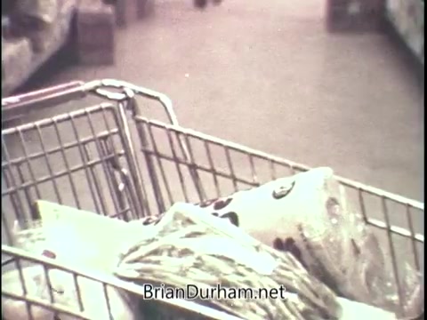 The still from the 1972 Saran Wrap Freshness Test commercial depicts a shopping cart filled with various bags, likely containing food items. The setting appears to be inside a grocery store, with shelves visible in the background. The overall color tone is soft, suggesting a vintage aesthetic typical of that era. The focus is on the cart, emphasizing the idea of preserving freshness with Saran Wrap.