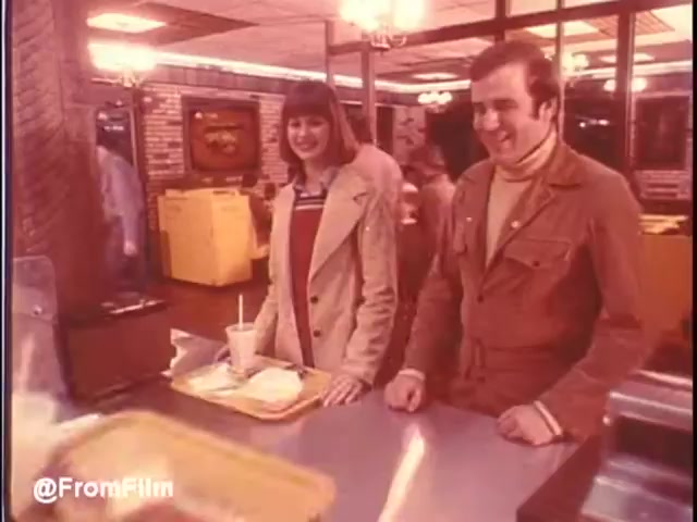 The still captures a moment inside a McDonald's in New Orleans from 1976. A young couple stands at the counter, looking pleased. The man is dressed in a brown outfit, while the woman sports a cozy, stylish look with a light-colored coat. They're in a classic McDonald's interior, featuring a warm ambiance with brick walls and a busy setting in the background. The scene evokes a sense of nostalgia for fast-food dining in the '70s. Would you eat at McDonald's in New Orleans? Maybe...   Follow us on Twitter @FromFilm.
