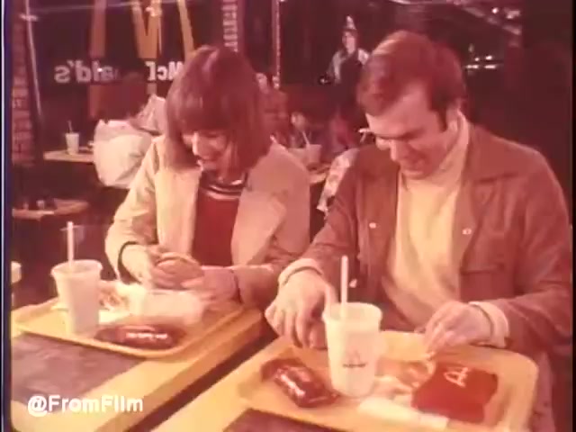 The still captures a cozy moment inside a McDonald's restaurant in New Orleans, circa 1976. A young couple sits at a bright yellow tray table, engrossed in their meal. The woman, wearing a light green jacket, seems excited as she unwraps her food, while the man, dressed in a brown jacket, looks content as he prepares to dig in. You can see iconic red and yellow McDonald's branding in the background, adding to the nostalgic atmosphere of the scene.   Would I eat at McDonald's in New Orleans? Maybe...   Follow us on Twitter @FromFilm.