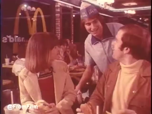 A lively 16mm still from a 1976 McDonald's commercial in New Orleans captures a warm moment inside the restaurant. A young employee, wearing a distinctive paper hat and a blue uniform, engages with a smiling couple at a table. The woman, dressed in a colorful sweater, looks intrigued, while the man in a brown jacket seems amused. The iconic golden arches logo and bustling restaurant atmosphere create a nostalgic vibe. Would I eat at McDonald's in New Orleans? Maybe...   Follow us on Twitter @FromFilm.