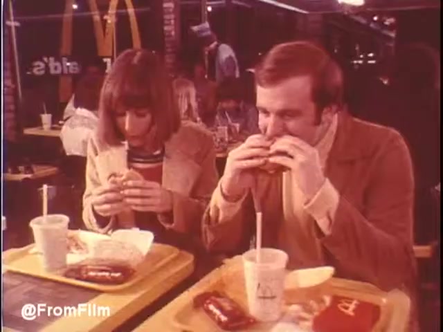 The still captures a moment from a 1976 McDonald's commercial set in New Orleans. In the frame, a man and a woman are seated at a bright yellow tray table, each holding a burger. The atmosphere is casual, with others in the background enjoying their meals. The interior showcases a classic McDonald's design of the era, hinting at a lively dining experience. The color palette reflects the 1970s style, giving the scene a nostalgic feel. Would you eat at McDonald's in New Orleans? Maybe...   Follow us on Twitter @FromFilm.