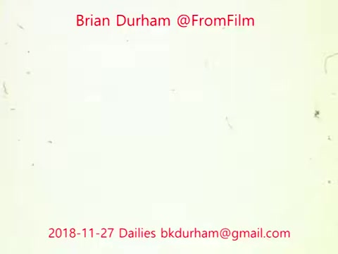 The still features a simple, clean background with the text prominently displayed. It includes the name 'Brian Durham' and the handle '@FromFilm,' along with the date '2018-11-27' and an email address. The overall appearance suggests a focus on film dailies, likely showcasing a moment from the production process, but the visual content is minimal. The background shows some grain and imperfections typical of 16mm film.