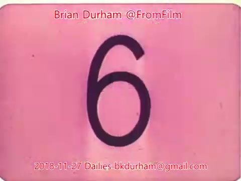 The still features a large black numeral '6' on a pink background, indicating a scene number for film editing. The image includes credits for Brian Durham and the date, November 27, 2018, along with a contact email related to the film dailies. The style suggests it is part of a film reel, likely during the editing process.