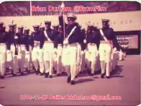 The still features a military parade, showcasing soldiers in uniform marching in formation. They are wearing ceremonial attire, which includes white trousers and jackets, with some bearing sashes. The scene appears to be a formal event, emphasizing discipline and ceremonial duties. The image's color and vintage quality suggest it is a historic film still from the 16mm format.