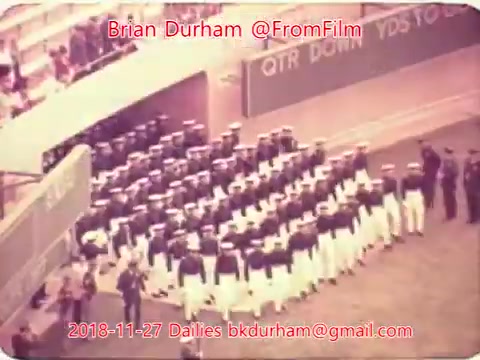 The still depicts a formation of individuals in military uniforms, likely in a ceremonial or parade setting. They are arranged in rows, facing a reviewing stand or audience. The backdrop includes signage indicative of a sports venue, suggesting a military event or presentation may be taking place. The color tone suggests it is an older 16mm film.