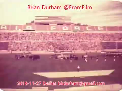 The image is a 16mm still showing a crowded stadium with the words 'AIR FORCE' prominently displayed in the background. The scene captures a sporting event or gathering, with fans in the stands and activities occurring on the field. The overall tone appears nostalgic, likely from an earlier era, given the film format. The date and contact information suggest it's part of a film archive or project.