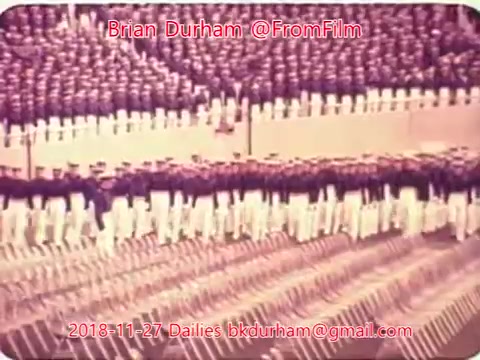 The still depicts a large group of marching individuals, likely in a military or ceremonial formation. The scene features a well-organized display, with participants in uniforms of varying colors and styles, creating a visual rhythm. The setting appears to be outdoors, possibly at a parade or official event, characterized by a large audience and staged rows. The overall tone of the image suggests a formal occasion.