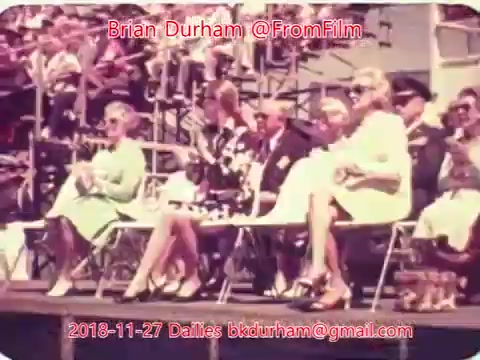 The still depicts a vintage scene featuring several well-dressed individuals seated in folding chairs, likely at an outdoor event. The setting appears lively, with a crowd visible in the background. The fashion suggests a mid-20th century context, emphasizing a formal or celebratory atmosphere.