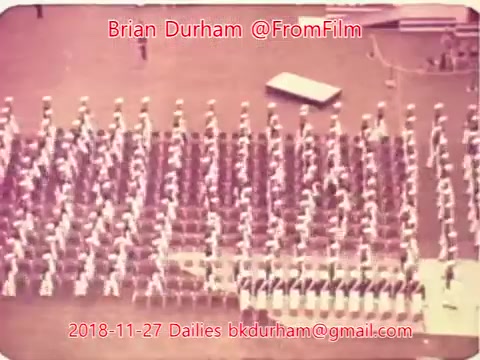 The still depicts a large group of uniformed individuals arranged in a precise formation, suggesting a military or marching band context. The scene is shot from a high angle, emphasizing the orderly layout of the participants. It has a vintage appearance, characteristic of 16mm film, with a soft focus and muted colors.