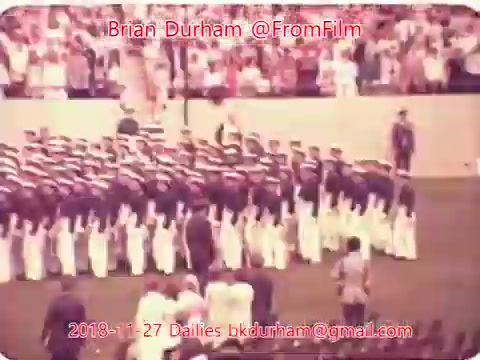 The still depicts a group of uniformed individuals, likely a military or ceremonial unit, standing in formation on a field. They are arranged in rows, with spectators visible in the background, indicating a formal event or gathering. The image has a vintage quality, characteristic of 16mm film.