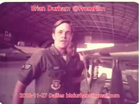 The still features a man in military uniform, standing beside aircraft in a hangar. He has a friendly expression and is looking at the camera. The environment is industrial, with visible planes in the background and a high ceiling. The image is likely from a film or documentation related to military aviation.