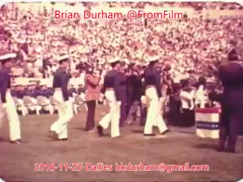 The still features a scene from a sports event, likely a football game, taking place in a stadium filled with a large crowd. A group of individuals in military uniforms appears to be participating in a ceremony or presentation on the field, while others look on. The atmosphere is lively, with spectators engaged in the event.