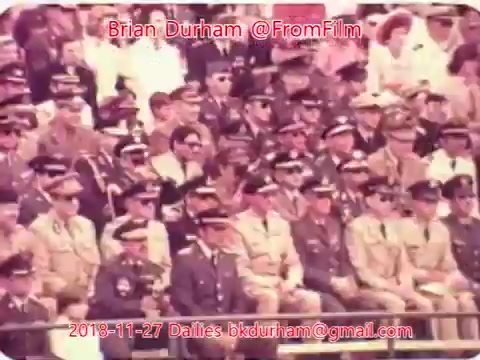 The still likely depicts a crowd of uniformed military or police personnel seated in a formal gathering, possibly a parade or ceremony. The image has a vintage quality characteristic of 16mm film, with a warm color palette. There are individuals wearing various ranks and uniforms, some with sunglasses, suggesting a structured and official atmosphere.
