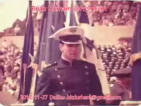 The still depicts a young man in a military uniform, wearing a white cap with a visor. He stands in front of a crowd, flanked by multiple flags, suggesting a ceremonial event or parade. The background shows numerous spectators, indicating a public occasion. The image has a vintage feel, characteristic of 16mm film.
