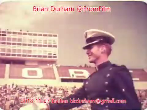 The still shows a smiling man in a military uniform and cap, likely a ceremonial figure, walking in a stadium filled with spectators. The background features seating and the prominent letters 'O R B.' The image has a vintage aesthetic, typical of 16mm film, suggesting a nostalgic or historical context.