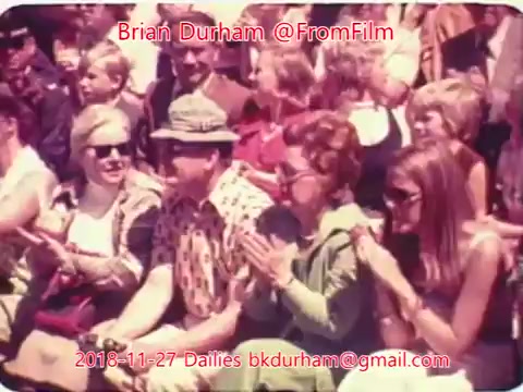 The still features a group of seated individuals, likely enjoying an event or performance outdoors. Some people are smiling and clapping, conveying a sense of community and enjoyment. The scene is brightly colored, typical of 16mm film aesthetics from that era. Notable fashion styles of the period, such as sunglasses and patterned clothing, are evident among the attendees.