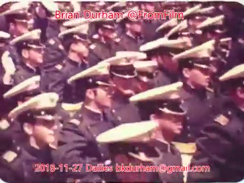 The still depicts a gathering of uniformed individuals, likely in a military or ceremonial context, sitting closely together. Their uniforms are characterized by distinct hats and insignia, suggesting a formal event. The focus is on the collective presence of the group, highlighting their disciplined demeanor and unity. The image appears to have a vintage quality, typical of 16mm film, with subdued colors.