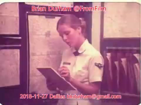 The still shows a woman in a uniform, possibly a nurse or an airline attendant, holding a clipboard and looking thoughtful as she writes. She is in an office or a workplace setting, with maps or charts visible in the background. The image is styled in a retro 16mm film aesthetic, indicative of the time period represented.