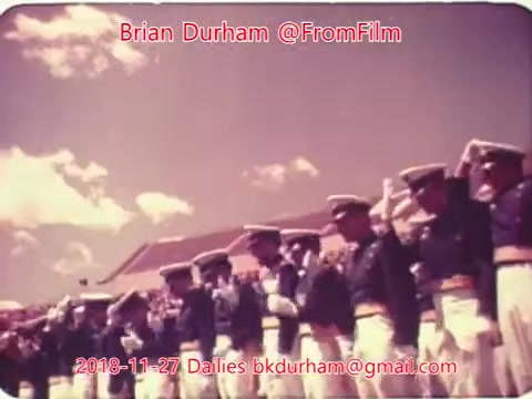 The still shows a group of uniformed military personnel standing in formation, saluting. They are dressed in formal attire, and the scene appears to be set outdoors under a cloudy sky. The color palette suggests a vintage feel, typical of 16mm film from that era. The image is branded with text indicating the source and date.