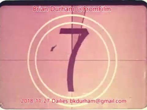 The still depicts a vintage film countdown graphic, showing the number '7' in a prominent position within concentric circles. The overall aesthetic reflects an old-school film style, typical of 16mm dailies. The image features a muted color palette, consistent with the era of the film. There are overlay details including the creator's name, date, and contact information.