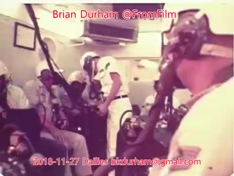 The still depicts a group of individuals in a confined space, possibly a command center or control room. Most of them are wearing helmets and oxygen masks, suggesting a focus on safety or a high-stakes environment. One person, dressed in white, appears to be engaged with the others, possibly giving instructions or information. The overall atmosphere suggests teamwork and preparation, likely in a scientific or experimental context.