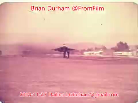The still from '@FromFilm Dailies November 27, 2018' depicts a jet aircraft taking off or landing on a runway, with smoke trailing behind. The background shows clear skies and some structures, possibly airport facilities. The image has a vintage appearance typical of 16mm film.