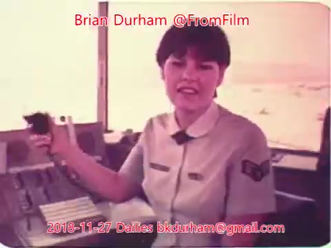 The still features a woman in a light-colored uniform, smiling and gesturing while seated at a control panel. The background suggests a workspace, possibly in an operational setting. The image has a vintage quality, typical of 16mm film, and includes identifying text with names and a date.
