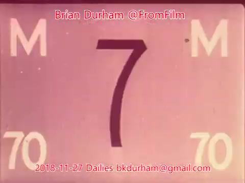 The still features a numbered frame from a 16mm film, displaying the number '7' prominently in the center, surrounded by 'M' on either side and the numbers '70' at the bottom. The background is a faded pink hue, lending a vintage aesthetic to the image. There are also text details indicating the creator, date, and contact information.