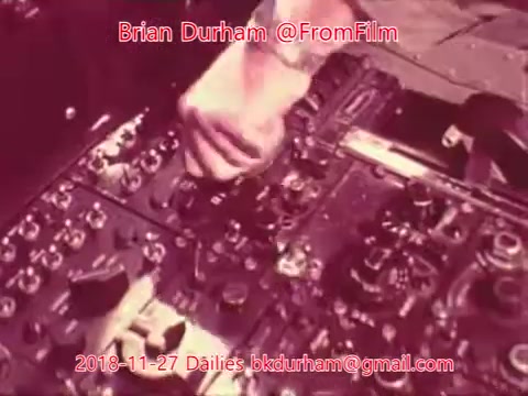 The still from the 16mm film shows a close-up of a person’s hand manipulating various controls on a panel. The panel is filled with knobs, switches, and dials, suggesting a technical or mechanical context. The color tones appear to be warm, likely due to the film's age or processing. The image conveys a sense of action or operation, reflecting a moment of engagement with equipment.