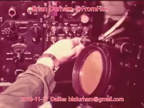 The still features a hand adjusting a dial or control on a vintage piece of equipment, likely related to film or audio technology. The background includes various knobs and switches, indicative of complex machinery. The colors appear faded, characteristic of older 16mm film. Superimposed text identifies the creator and date of the still.