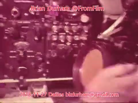 The still features a close-up of a control panel with various knobs and switches, suggesting a technical or engineering context. In the foreground, a hand is seen holding a pair of headphones. The overall color tone has a vintage quality, indicating it was shot on 16mm film. Text overlays provide details about the date and source of the image.
