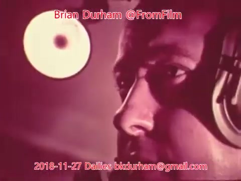 The still features a close-up of a man's face, captured in a warm, muted color palette. He appears focused, likely engaged in a recording or production process, with headphones on and a soft light source illuminating him. The image suggests a behind-the-scenes moment from a film or audio project, reflecting a sense of concentration and creativity.