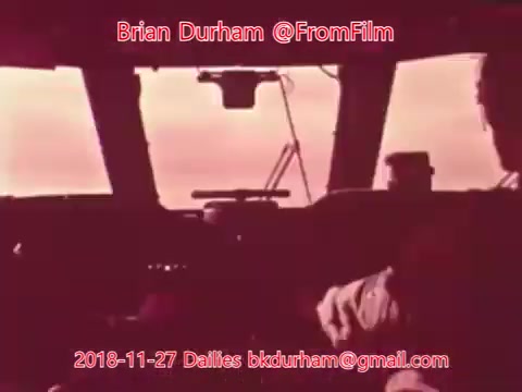 The still likely captures a cockpit or control room scene, featuring dim, red-toned lighting. The image shows figures possibly engaged in piloting or observation, with instruments and controls visible. There’s an emphasis on the atmosphere created by the color scheme, contributing to a moody or tense feel.