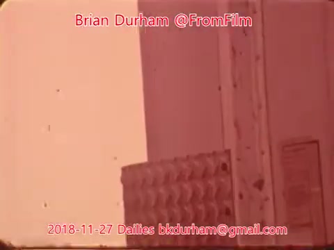 The still features a close-up view of a portion of what appears to be a textured surface, possibly a wall or a machine, with visible wear and tear. A label or instruction panel is present but not clearly readable. The overall color tone is warm, likely due to the film's aging or processing, contributing to a nostalgic feel. Text overlay indicates the date and credits related to the film's production.